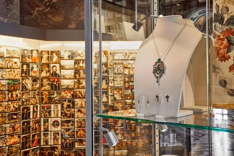 A display case featuring a necklace with a green stone and matching earrings is shown in the foreground. The background reveals a wall filled with numerous photographs, creating a vibrant and artistic atmosphere. The focus is on the jewelry set prominently featured.