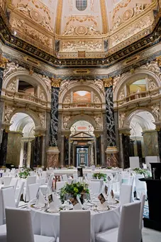 Ein eleganter Veranstaltungsraum mit runden Tischen, die mit weißen Tischdecken und Blumenarrangements dekoriert sind. Die Wände sind kunstvoll verziert, mit hohen Säulen und einem beeindruckenden Deckenmuster. Der Raum strahlt eine festliche Atmosphäre aus, geeignet für ein gehobenes Event oder eine Feier.