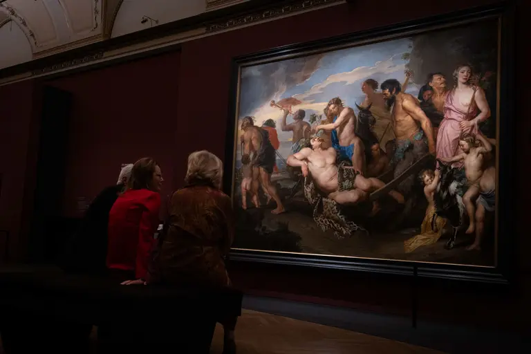 Drei Personen sitzen vor einem großen Gemälde, das eine mythologische Szene mit vielen Figuren zeigt. Die Figuren sind in lebhaften Posen dargestellt und scheinen eine dramatische Handlung zu vollziehen, während der Hintergrund eine wolkige Landschaft zeigt.