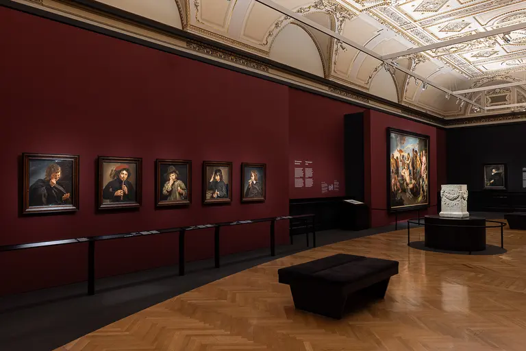 Das Bild zeigt einen Ausstellungsraum mit Gemälden an einer rot gestrichenen Wand. Zu sehen sind mehrere Porträts in runden Rahmen sowie ein großes Gruppenbild auf der rechten Seite. Der Boden ist aus Holz, und in der Mitte steht eine schwarze Sitzbank.