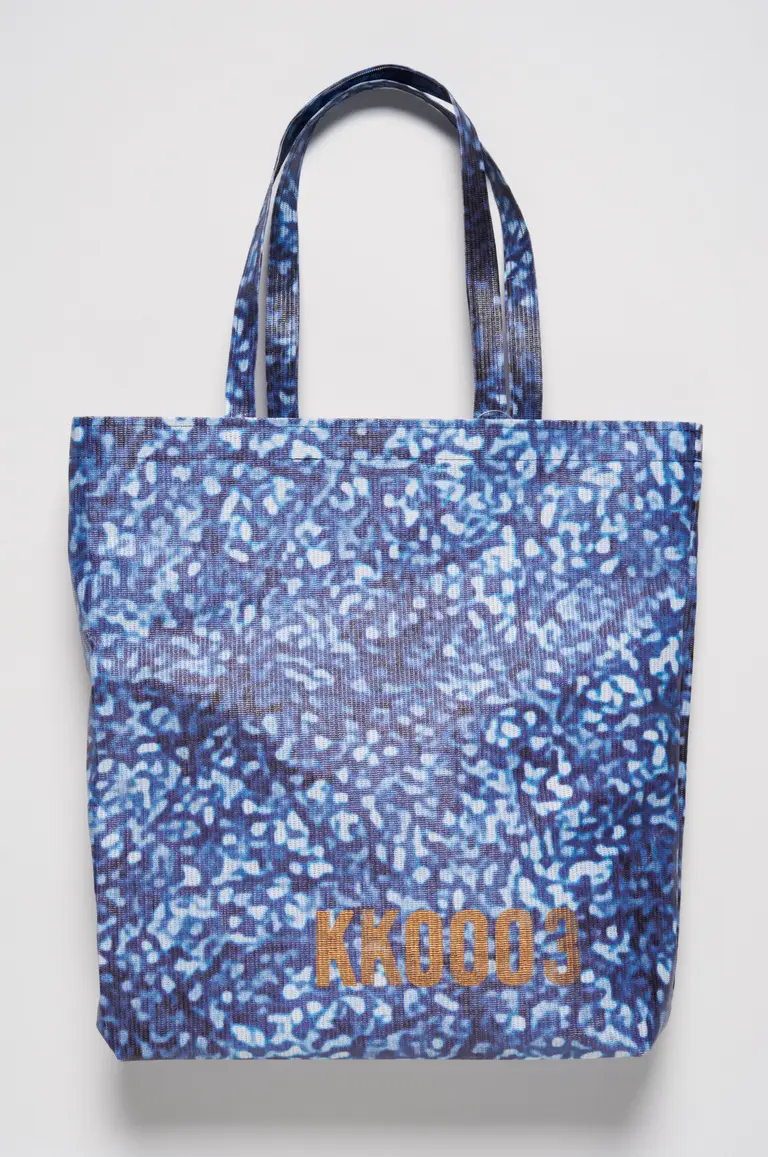 Eine blaue Tragetasche mit einem strukturierten Design aus verschiedenen Blautönen. In der unteren Ecke befindet sich die Aufschrift