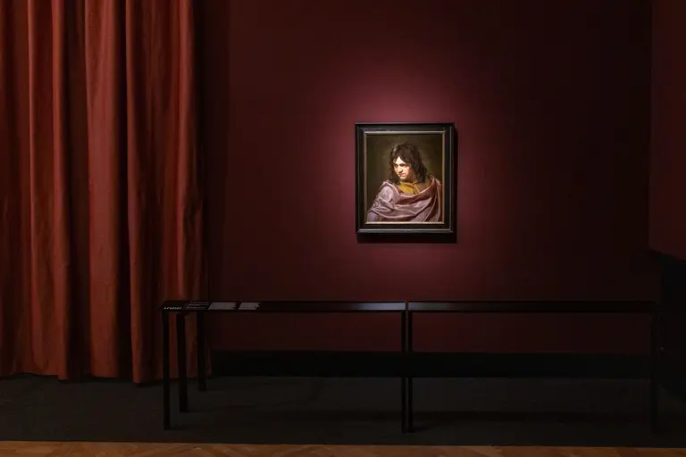 Ein Porträt einer Frau mit dunklen Haaren, die einen lila Umhang trägt, ist an einer Wand einer Galerie beleuchtet. Der Hintergrund ist rot, und ein schwarzes Exhibitionstisch ist im Vordergrund sichtbar. Vor dem Gemälde gibt es keine weiteren störenden Elemente.