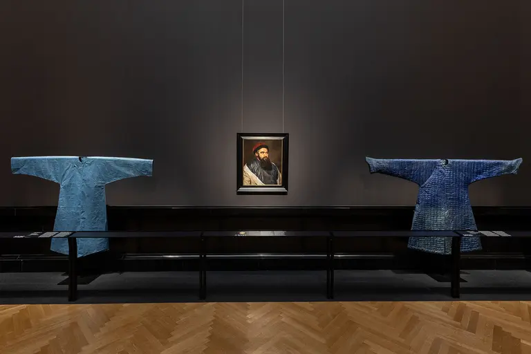 Im Bild sind zwei blaue traditionelle Gewänder auf einem Tisch zu sehen, die symmetrisch angeordnet sind. In der Mitte hängt ein Porträt eines Mannes mit Bart und Hut an der Wand. Der Hintergrund ist dunkel, was die Kleidungsstücke und das Porträt hervorhebt.