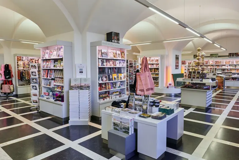 A spacious bookstore featuring neatly organized shelves filled with magazines and books. The interior is brightly lit with high ceilings and a checkerboard floor pattern, creating an inviting atmosphere for readers and visitors.