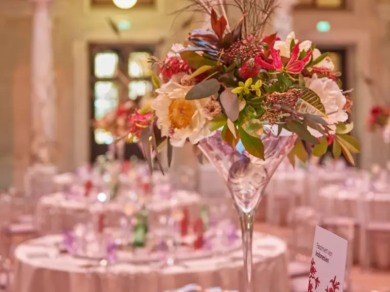 Ein elegantes Blumenarrangement in einer hohen Glasvase mit Pfauenfedern ziert einen Tisch in einem festlich dekorierten Raum. Im Hintergrund sind gedeckte Tische mit buntem Geschirr und Materialien sichtbar, die eine festliche Atmosphäre schaffen.
