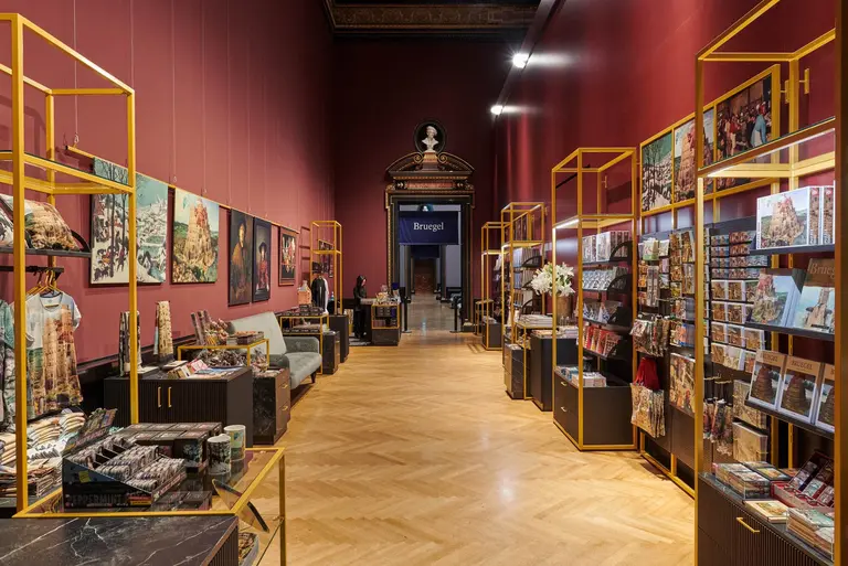 The image shows an art gallery with a spacious layout featuring golden display stands. On the walls are various framed artworks, while merchandise and books are presented on tables and shelves. The gallery has a rich red background, creating an inviting atmosphere for visitors.