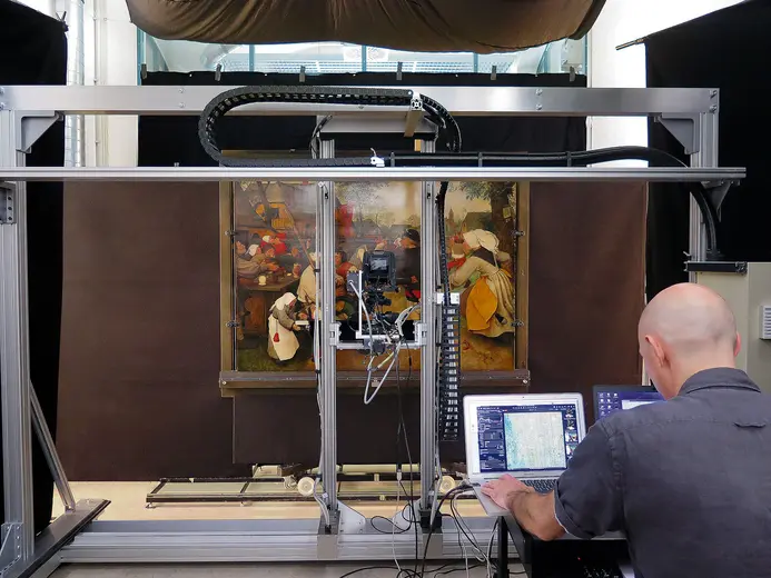 Ein Kunstwerk wird digitalisiert. Im Vordergrund sitzt eine Person an einem Computer, während eine Maschine das Gemälde erfasst. Der Hintergrund ist dunkel, um die Aufmerksamkeit auf das Kunstwerk zu lenken.