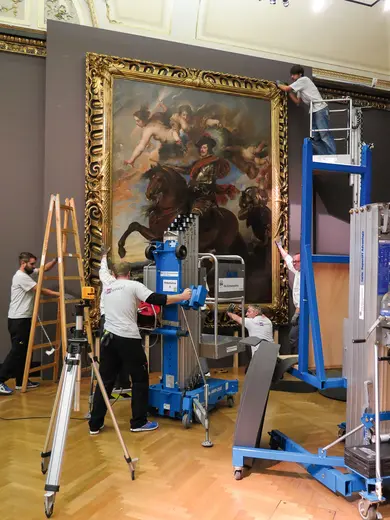 Fünf Personen arbeiten daran, ein großes Gemälde in einem Ausstellungsraum aufzuhängen. Zwei Personen stehen auf Leitern, während andere eine Hebebühne und Werkzeuge benutzen, um das Kunstwerk an der Wand zu sichern. Der Rahmen des Gemäldes ist reich verziert.