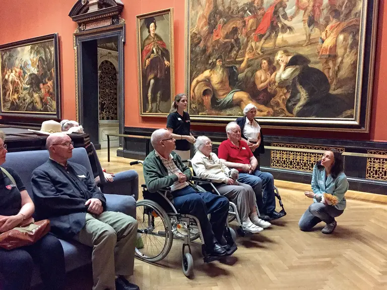 A group of elderly visitors, some seated in wheelchairs, listens attentively as a guide explains artwork in a museum. The room features large paintings on the walls, and the ambiance is warm and inviting, encouraging engagement with the art.