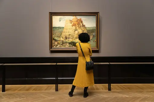 A person with curly hair, wearing a yellow dress, stands in front of a large painting depicting a tower with a complex structure, likely representing the Tower of Babel. The setting is an art gallery with wooden floors and a neutral-colored wall.