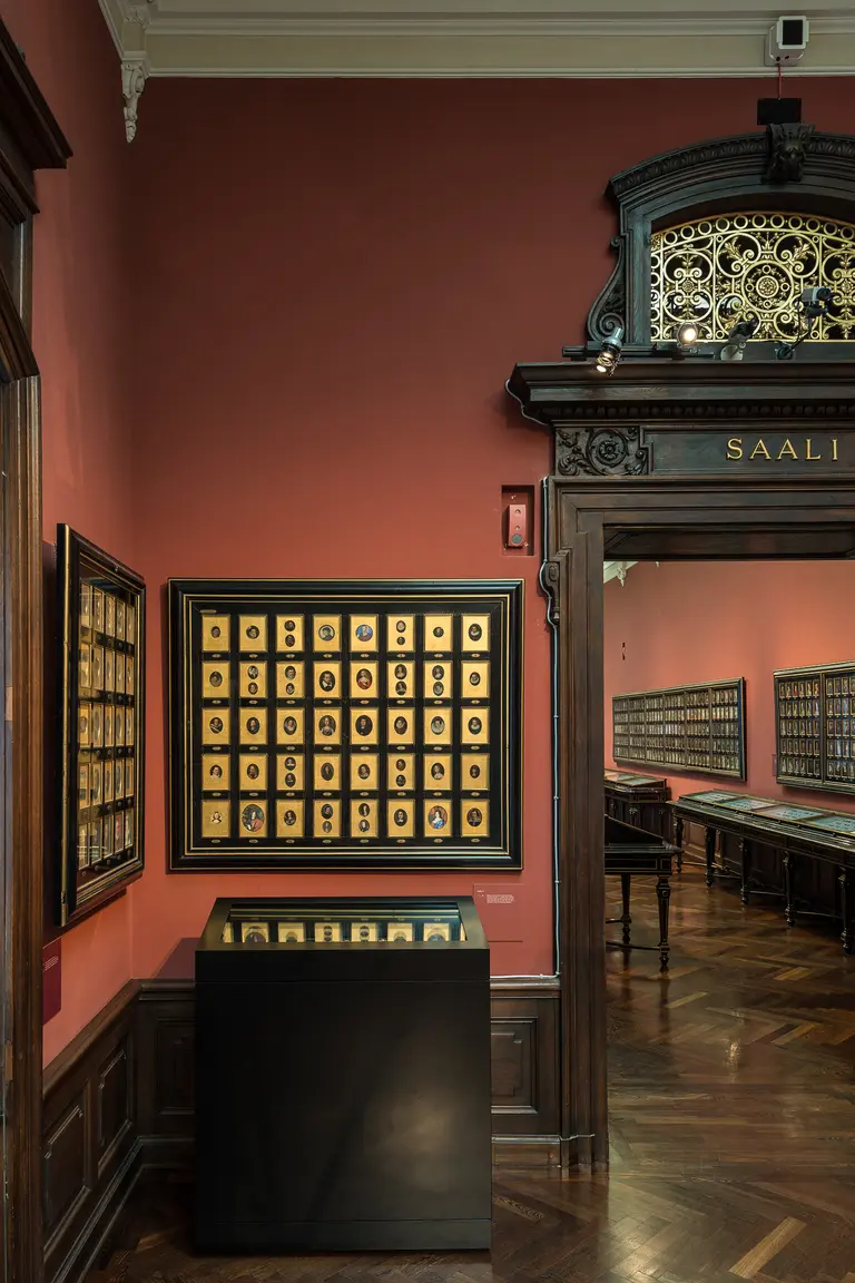 In einem Ausstellungsraum mit roter Wand sind mehrere Bilderrahmen zu sehen, die eine Vielzahl von Fotografien in goldenen Rahmen zeigen. In der Mitte des Raumes befindet sich ein schwarzer Tisch mit weiteren Exponaten. Der Eingang zu einem weiteren Raum ist gut sichtbar.