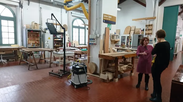 Eine Holzwerkstatt mit mehreren Tischen und Regalen. Zwei Personen unterhalten sich; eine trägt ein lila Kleid und die andere ist in Schwarz gekleidet. Im Raum sind verschiedene Werkzeuge und Materialien für die Holzverarbeitung sichtbar. Es herrscht eine helle und einladende Atmosphäre.