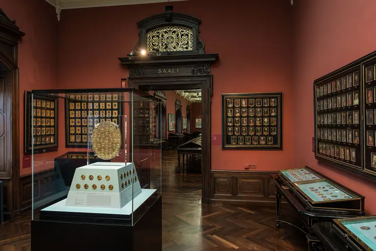 Im Bild ist ein Ausstellungsraum zu sehen, der mit roten Wänden und Holzböden gestaltet ist. In der Mitte steht ein Glasdisplay, das ein goldenes Objekt präsentiert. Im Hintergrund sind Wände mit gerahmten Bildern zu sehen, die einen historischen Kontext suggerieren.