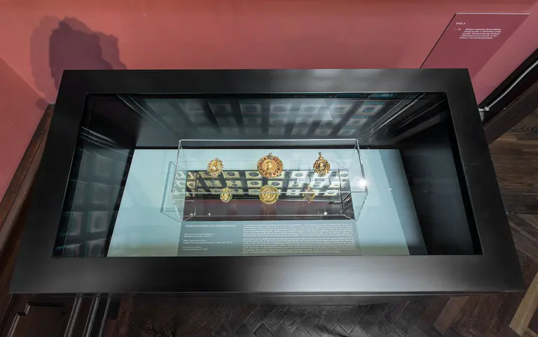 In der Abbildung ist eine Vitrine zu sehen, die verschiedene kunstvolle goldene Schmuckstücke zeigt. Diese sind auf einem weißen Hintergrund platziert, in einem eleganten, modernen Ausstellungsdesign. Die Vitrine befindet sich in einem Museum mit dunklem Holzboden.