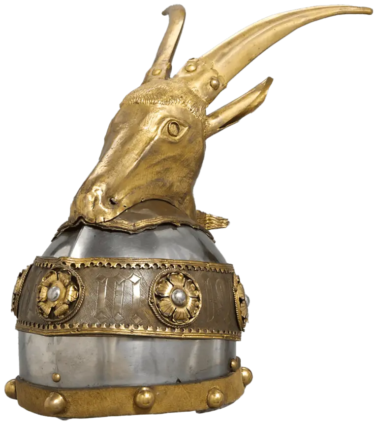 A decorative helmet features a stylized goat head made of gold. The helmet's base is metallic with engraved patterns and adorned with circular gold ornaments and rivets, emphasizing its historical and artistic significance.
