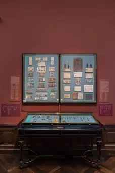Das Bild zeigt ein Ausstellungsstück in einem Museum. An der Wand befinden sich zwei große Tafeln mit verschiedenen alten Geldscheinen und Dokumenten. Vor der Wand steht ein Ausstellungstisch mit Münzen und Schmuck, die ebenfalls zur Sammlung gehören.