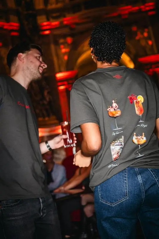 Two people are socializing in a lively setting, wearing gray t-shirts featuring illustrations of cocktails. One person holds a drink, while the other is facing away, showcasing the drink designs on their shirt. The background has warm lighting, suggesting a festive atmosphere.