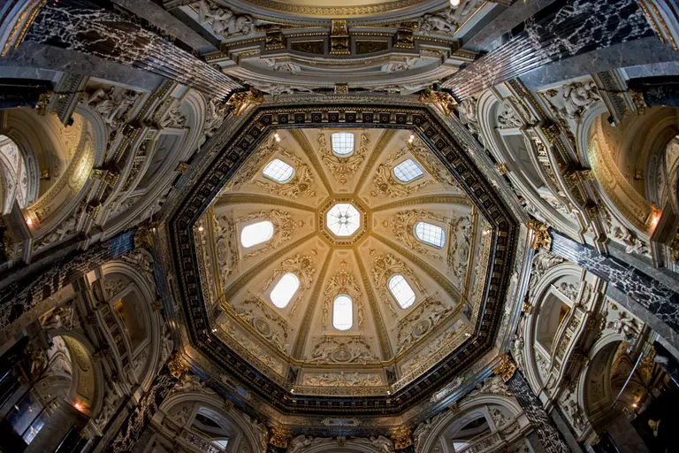 The image captures the intricate ceiling of an octagonal dome, featuring ornate decorations and several windows allowing natural light to filter through. The design displays a combination of architectural elegance and artistic detailing, drawing the viewer's eye upward toward the center.