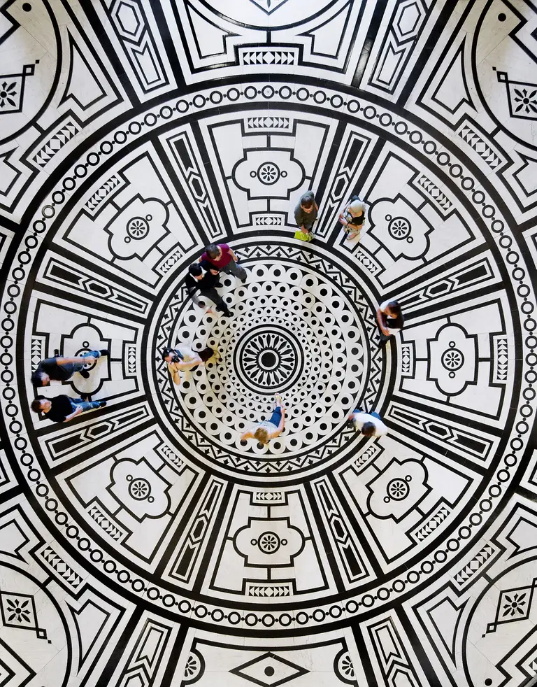 Ein Rundmuster in Schwarz-Weiß auf dem Boden im KHM ist zu sehen. Mehrere Personen stehen und bewegen sich innerhalb dieses Musters. Das Design ist geometrisch und symmetrisch, mit auffälligen Kreisen und floralen Motiven im Mittelpunkt.