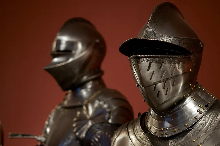 Zwei mittelalterliche Rüstungen stehen nebeneinander vor einem roten Hintergrund. Die glänzenden Metalloberflächen sind kunstvoll verziert, mit unterschiedlichen Helmarten, die die Gesichter der Rüstungen verdecken. Diese Rüstungen repräsentieren die Rüstkunst und den militärischen Stil vergangener Zeiten.