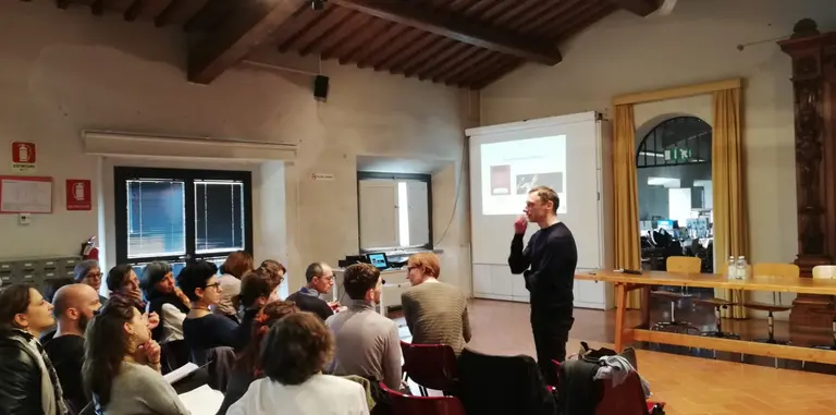 Ein Referent steht vor einem Publikum in einem Raum, während er spricht. Im Hintergrund ist ein Projektor mit Präsentationsinhalten sichtbar. Die Zuhörer sitzen in Reihen und schauen aufmerksam in die Richtung des Sprechers.