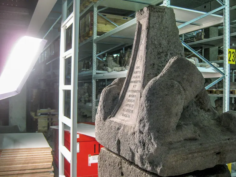 Ein antiker Stein mit Inschrift, der möglicherweise ein Denkmal oder eine Grabstätte darstellt. Der Stein hat eine dekorative Form und zeigt eine geprägte Schrift. Im Hintergrund sind Regale mit weiteren Objekten zu sehen, die in einem Lagerraum aufbewahrt werden.