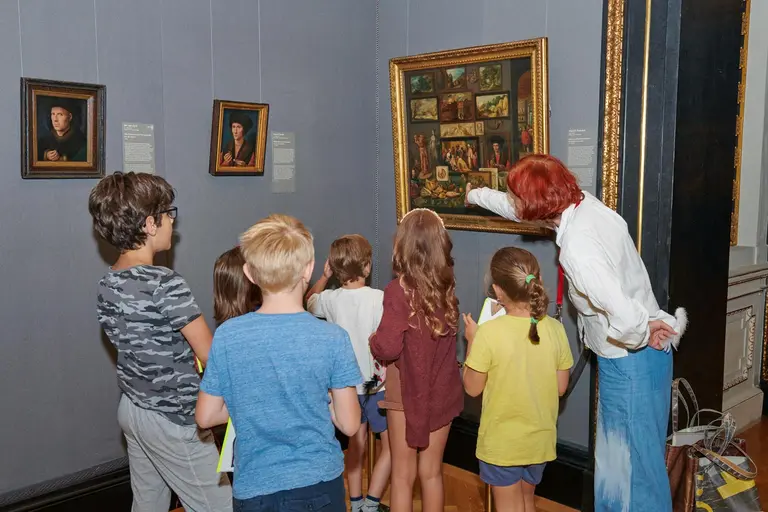 Eine Gruppe von Kindern steht vor einem Gemälde in einem Museum. Ein Erwachsener zeigt auf das Bild und erklärt etwas. Im Hintergrund sind weitere Bilder an der Wand zu sehen. Die Kinder erscheinen aufmerksam und interessiert.