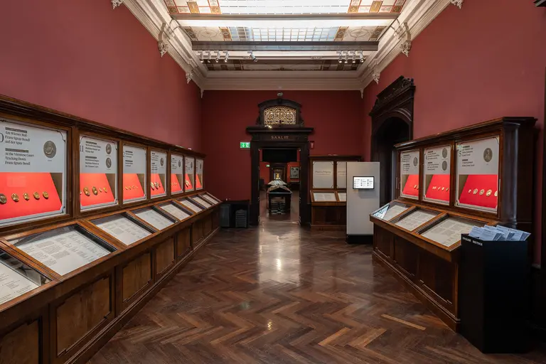 Das Bild zeigt einen Ausstellungsraum mit Holzmöbeln und Vitrinen. In den Vitrinen sind Münzen ausgestellt, die auf roten Unterlagen präsentiert werden. Der Raum hat hohe Decken und eine elegante Gesamtausstattung, die eine historische Atmosphäre schafft.