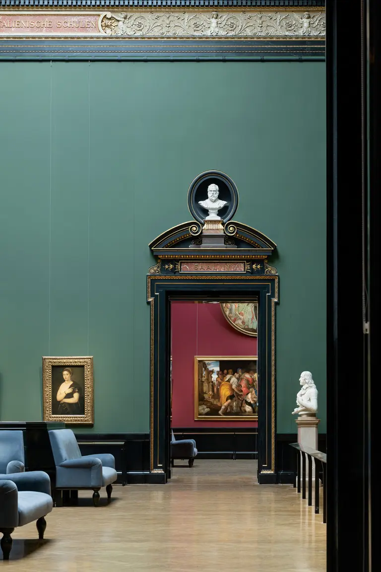 Ein lichtdurchfluteter Ausstellungsraum mit grünen Wänden. Im Mittelpunkt steht ein eleganter Türbogen, über dem eine Büste zu sehen ist. Links hängt ein Porträt, während im hinteren Bereich ein weiteres Gemälde zu erkennen ist. Gemütliche blaue Sessel laden zum Verweilen ein.