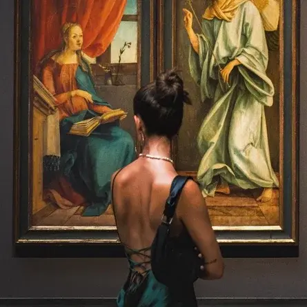 A woman stands in an art gallery, admiring two large paintings. The left painting features an angel visiting a seated figure, while the right painting depicts a vibrant scene with multiple people among nature. The gallery has a serene atmosphere, enhancing the viewing experience.