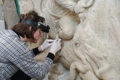 A woman wearing a magnifying visor and gloves carefully examines and works on a textured sculpture. She is focused on a section of the artwork, highlighting the detail and precision involved in the conservation process.