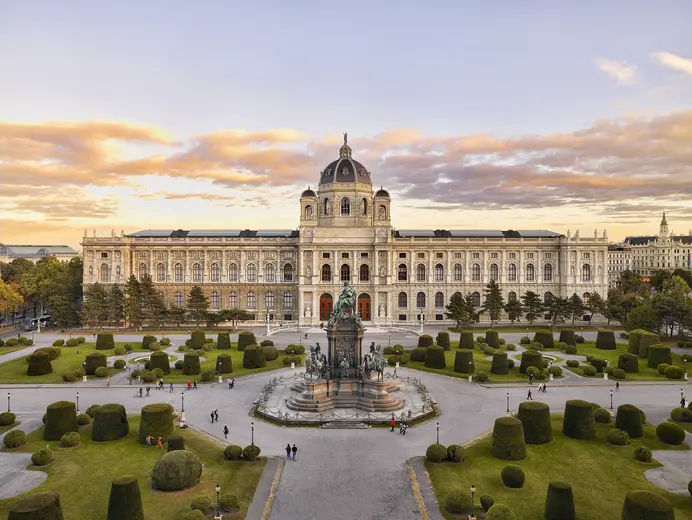 Das Bild zeigt das prächtige Kunsthistorische Museum in Wien, umgeben von gepflegten Gärten mit geometrisch geschnittenen Bäumen. Im Vordergrund steht eine Statue, und Besucher schlendern durch die Anlage, während der Himmel in warmen Farben leuchtet.