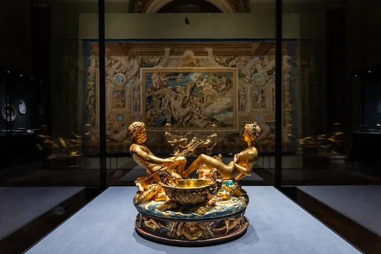 Eine goldene Skulptur zeigt zwei Figuren in einer Badewanne, die von kunstvollen Verzierungen umgeben sind. Im Hintergrund ist ein großes Wandgemälde zu sehen, das eine mythologische Szene darstellt. Die Präsentation erfolgt in einem Münzmuseum.