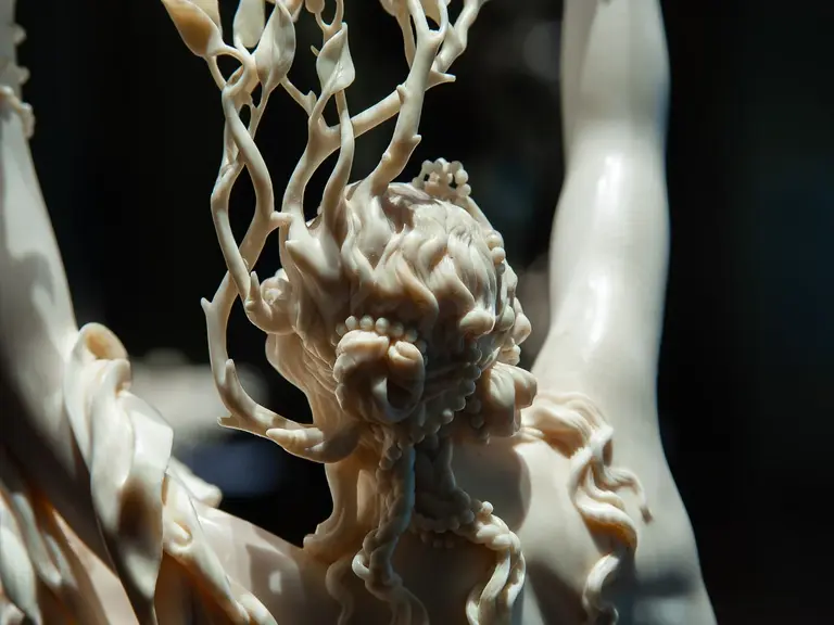 Eine detaillierte Skulptur zeigt eine weibliche Figur mit langen, fließenden Haaren, aus denen Äste und Blätter sprießen. Die Pose vermittelt Bewegung und Ausdruckskraft, während die Verarbeitung des Materials elegantes Licht und Schatten erzeugt.