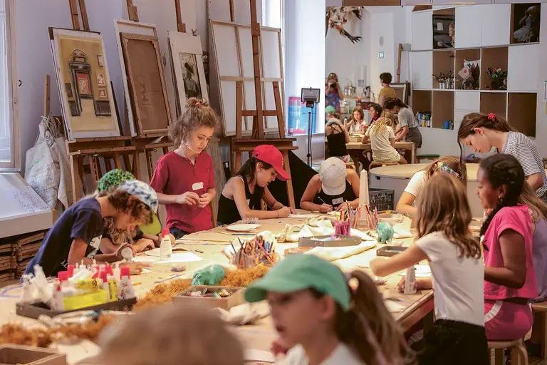 Eine Gruppe von Kindern arbeitet in einem kreativen Atelier. Sie sitzen an Tischen, zeichnen und basteln mit verschiedenen Materialien wie Farben und Pinsel. Im Hintergrund stehen Staffeleien und ein freundlicher, kreativer Raum ist sichtbar. Die Atmosphäre ist lebhaft und inspirierend.