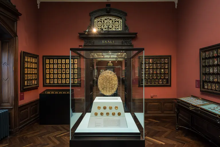 Das Bild zeigt einen Ausstellungsraum mit einem zentralen Vitrinenobjekt: einem runden Schild. Um die Vitrine sind kleinere Wandtafeln mit weiteren Objekten angeordnet. Die Wand ist in einem warmen Rotton gehalten, und die Atmosphäre wirkt historisch und elegant.