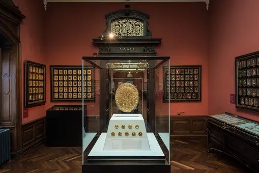 Das Bild zeigt einen Ausstellungsraum mit einem zentralen Vitrinenobjekt: einem runden Schild. Um die Vitrine sind kleinere Wandtafeln mit weiteren Objekten angeordnet. Die Wand ist in einem warmen Rotton gehalten, und die Atmosphäre wirkt historisch und elegant.