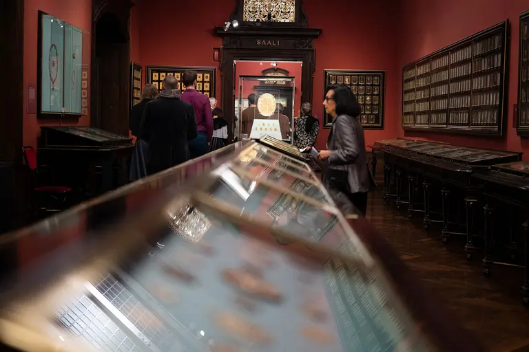 Das Bild zeigt einen Ausstellungsraum mit roten Wänden und Vitrinen, die verschiedene Ausstellungsstücke präsentieren. Besucher stehen um die Vitrinen herum und betrachten die Exponate. Am Ende des Raumes ist eine weitere große Vitrine zu sehen.