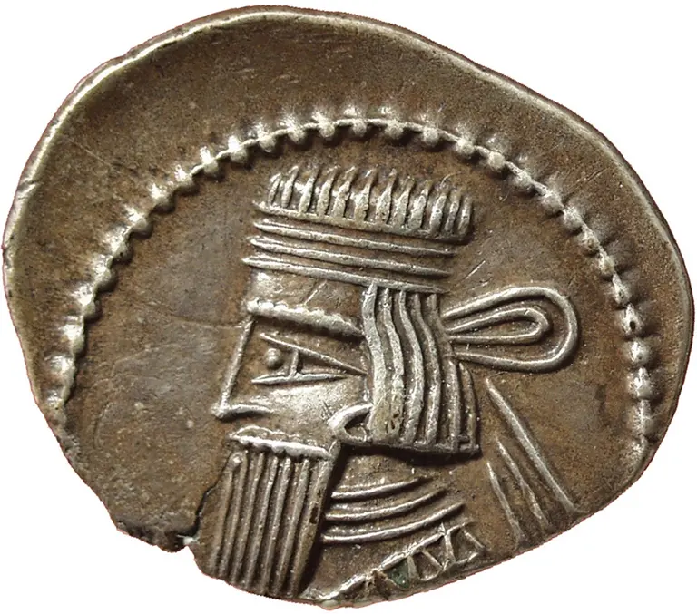 Eine antike Silbermünze, die ein menschliches Profil zeigt. Der Kopf hat eine lange, geflochtene Frisur und einen Bart. Vertiefungen und Linien auf der Münze deuten auf künstlerische Details hin, die die Gesichtszüge betonen. Die Münze hat eine abgerundete Form mit einer unebenen Kante.