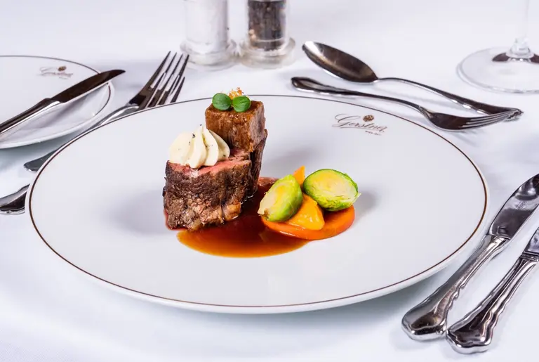 A gourmet dish featuring two pieces of beef, one topped with creamy mashed potatoes and garnished with green herbs. Served alongside colorful vegetables, including Brussels sprouts and orange segments, on a white plate. The elegant table setting includes polished cutlery and additional plates in the background.