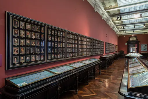 Das Bild zeigt einen Museumssaal mit einer langen Wand, die mit vielen Portraits in Rahmen bedeckt ist. Auf Tischen im Vordergrund sind Vitrinen mit verschiedenen Objekten ausgestellt. Der Raum hat eine elegante Holzfußboden und eine helle Decke mit Fenstern.