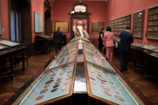 In einem Museum sind Besucher zu sehen, die um lange Vitrinen mit verschiedenen Münzen herumstehen. Der Raum hat orange Wände und eine elegante Holzdecke. Die Exponate sind sorgfältig ausgestellt, wodurch das Interesse der Besucher geweckt wird.