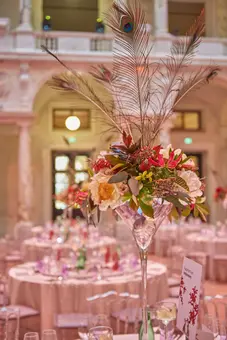 A tall, elegant floral arrangement featuring colorful flowers and peacock feathers sits in a crystal vase at the center of a beautifully set dining table, surrounded by chair backs. The backdrop hints at an ornate venue, creating an atmosphere suitable for a sophisticated event.