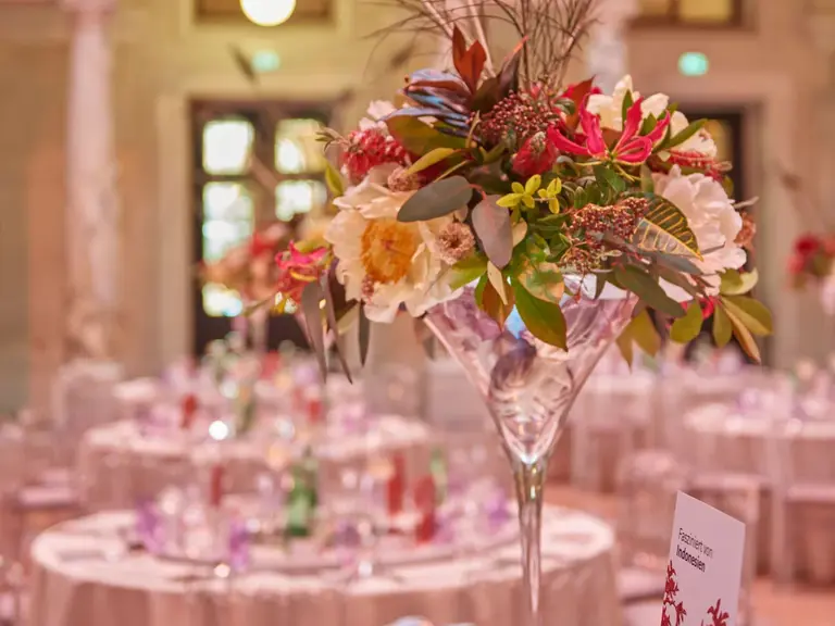 A tall, elegant floral arrangement featuring colorful flowers and peacock feathers sits in a crystal vase at the center of a beautifully set dining table, surrounded by chair backs. The backdrop hints at an ornate venue, creating an atmosphere suitable for a sophisticated event.