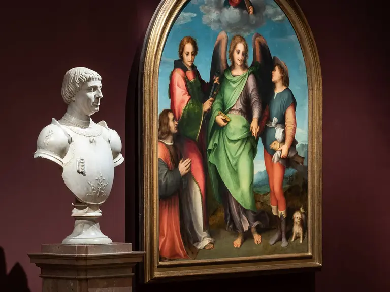 In einem Museum steht eine weiße Büste auf einem Sockel neben einem Gemälde. Das Gemälde zeigt vier Heilige, darunter einen Engel, umgeben von Himmel und Landschaft. Die Wände sind dunkelrot, und der Boden ist aus Holz.