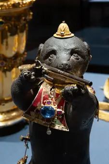 Eine kunstvolle Skulptur eines Bären, der ein Gewehr hält. Der Bär trägt eine prächtige Robe mit einem großen blauen Edelstein und einer goldenen Krone auf dem Kopf. Die Figur ist reich verziert und zeigt handwerkliches Können.