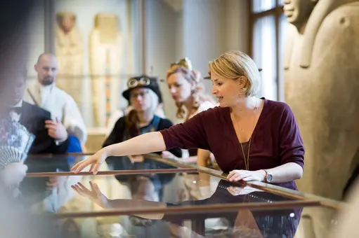 A museum guide engages a small group of visitors, gesturing toward an exhibit displayed under glass. The focus is on the guide’s hand as she points, while the audience listens attentively. The setting suggests a historical or cultural exhibit.