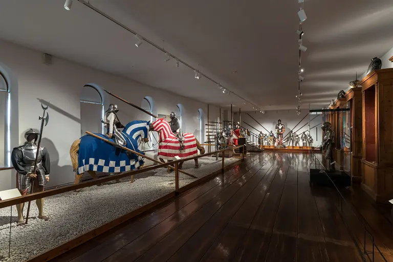 The image shows an exhibit in a museum featuring several armored knights on horseback, adorned with colorful banners. The room has wooden flooring and showcases various medieval weapons and armor, creating an atmosphere of historical significance.