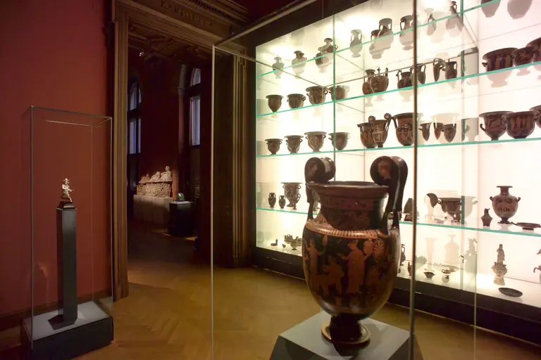 Das Bild zeigt eine Ausstellung mit antiken Töpferwaren in einem Museum. In einem gläsernen Display sind mehrere dekorierte Vasen und Krüge angeordnet. Im Vordergrund steht eine große, auffällige Vase, während im Hintergrund eine Statue auf einer Säule zu sehen ist.