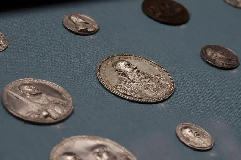 Eine Sammlung von silbernen Medaillen liegt auf einem blauen Hintergrund. Die Medaillen zeigen verschiedene Porträts, darunter eine größere Medaille mit einem bärtigen Mann. Die Anordnung der Medaillen ist unregelmäßig und es sind mehrere kleine Medaillen zu sehen.